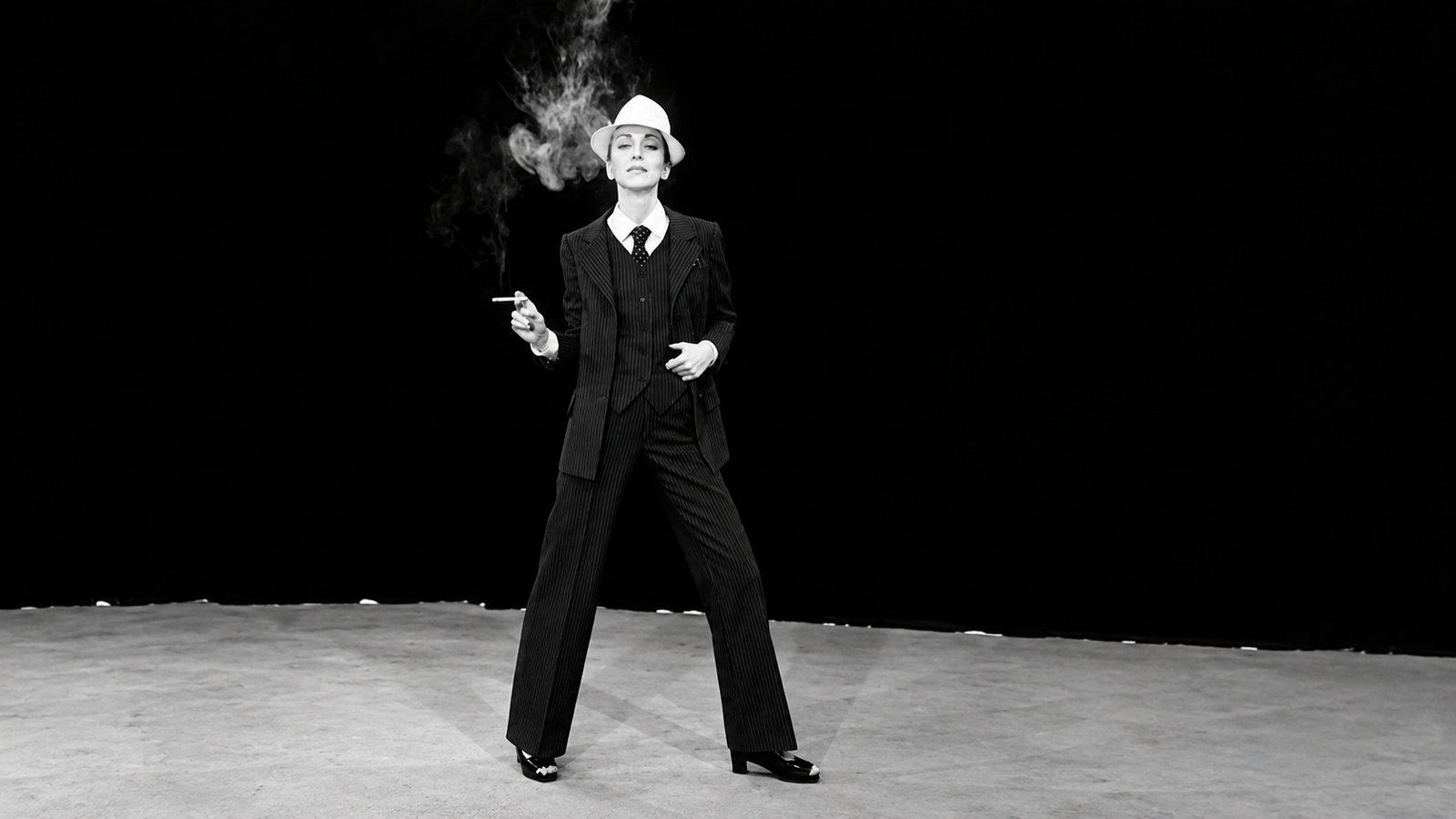 A black and white image of a woman in a dark suit, tie, and light fedora, holding a smoking cigarette.
