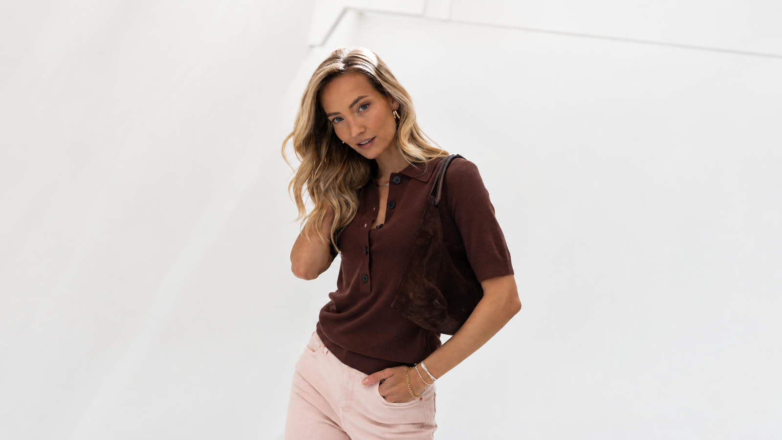 Eine Frau in einem braunen Hemd mit Kragen und einer rosa Hose hält eine dunkelbraune Tasche in der Hand und posiert vor einem weißen Hintergrund.