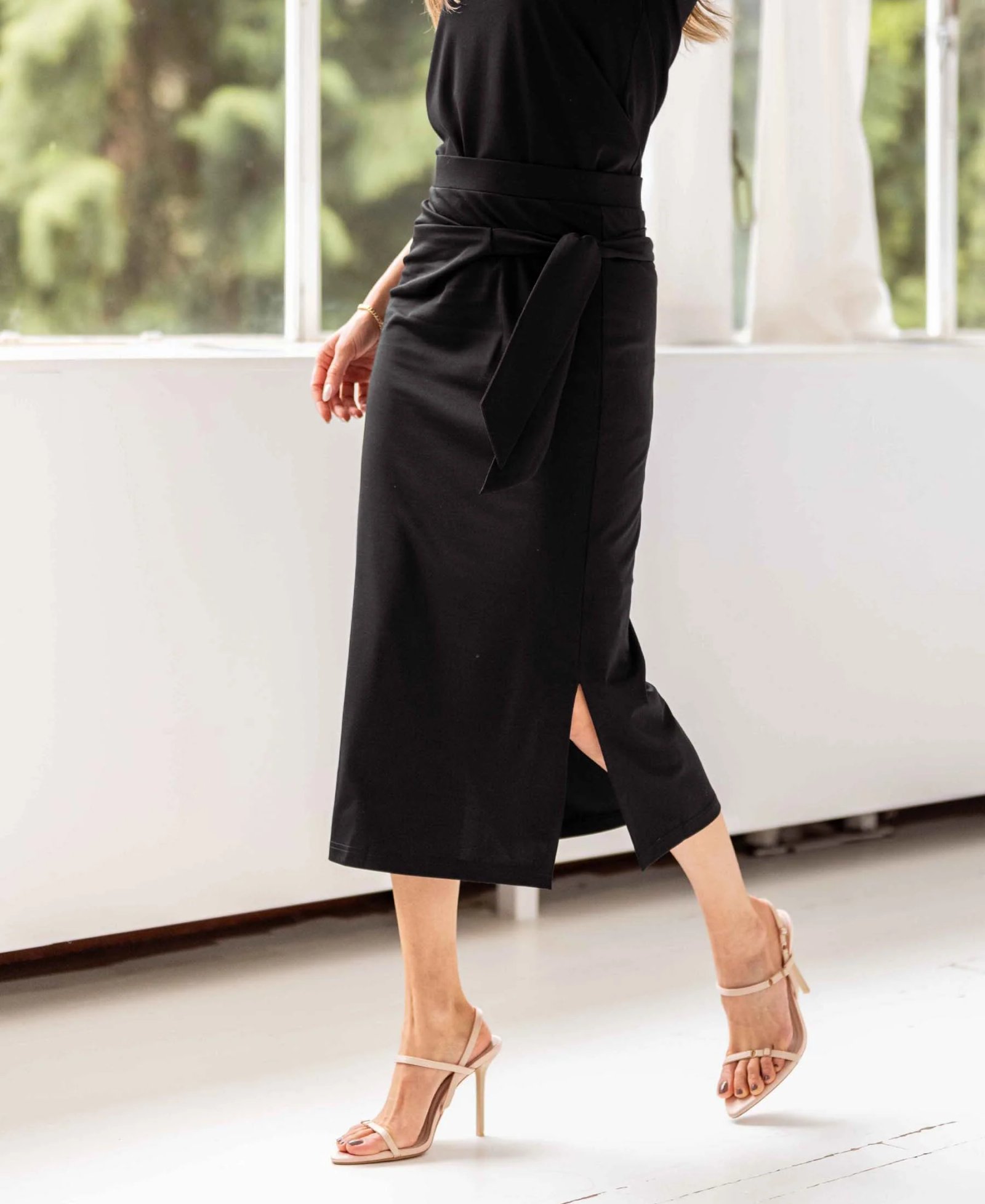 A person walks in a black tie-front midi skirt with a slit and nude strappy heels.