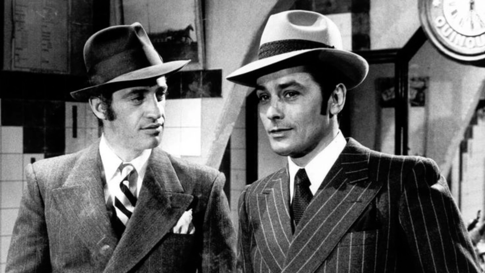 Schwarz-Weiß-Foto von Jean-Paul Belmondo und Alain Delon in Anzügen und mit Fedora-Hüten.