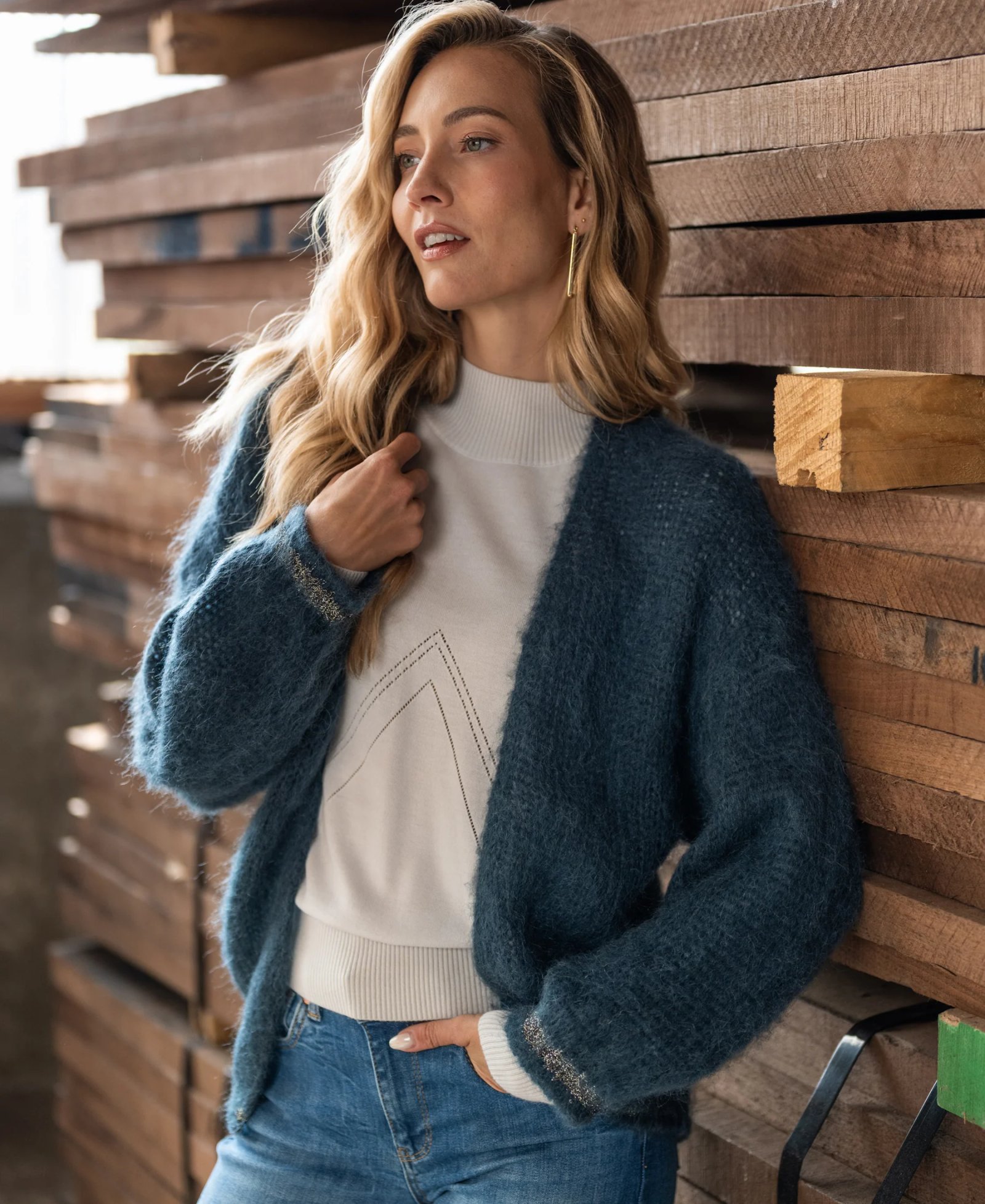 A woman with long blonde hair is wearing a teal, fluffy cardigan over a white sweater and jeans, leaning against a stack of wooden planks.