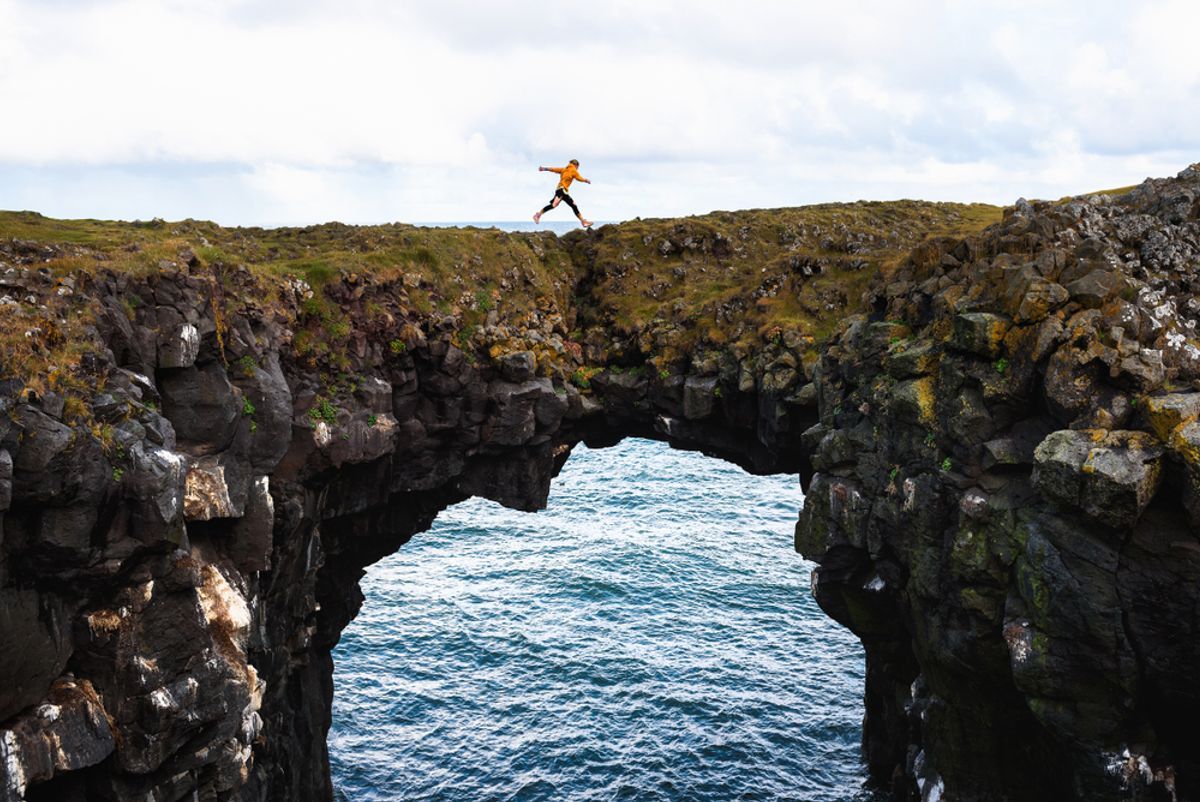Tourist jumps over a natural rock bridge connecting basalt cliffs in Arnarstapi, Iceland
