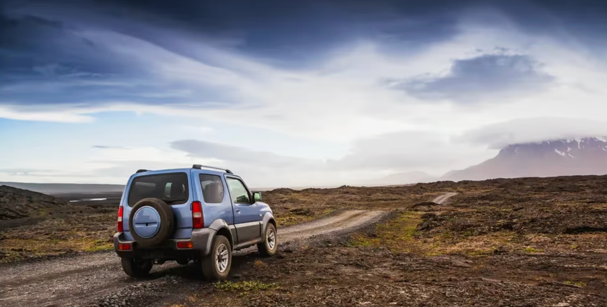 A blue 4x4 parked on a gravel road crossing a lava plain under a dramatic sky