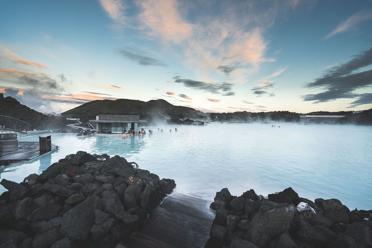 Bathers in the milky blue water of the Blue Lagoon at sunset, with black lava rocks in the foreground