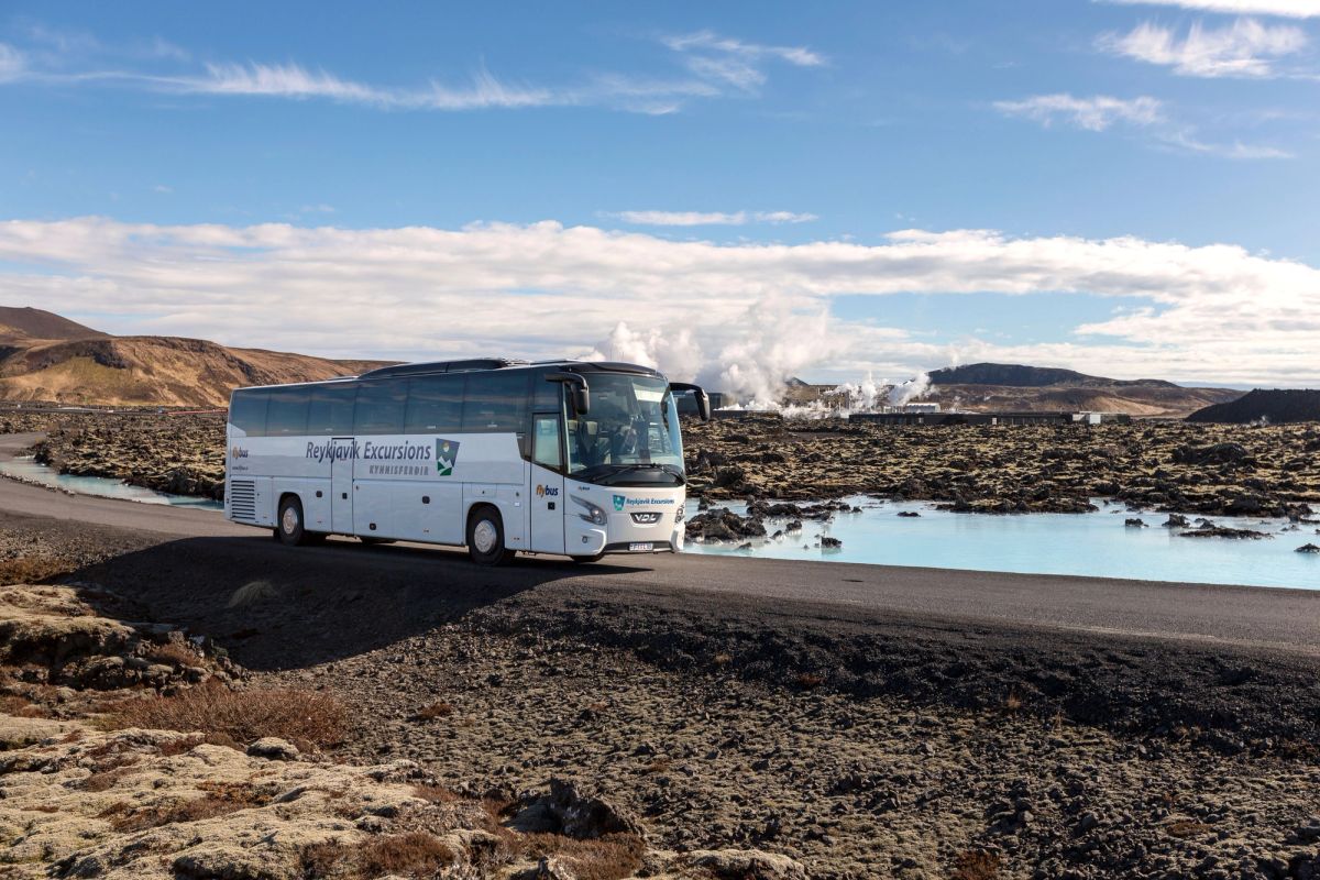 A tour coach on a road through a lava field, with steam rising from a geothermal plant behind