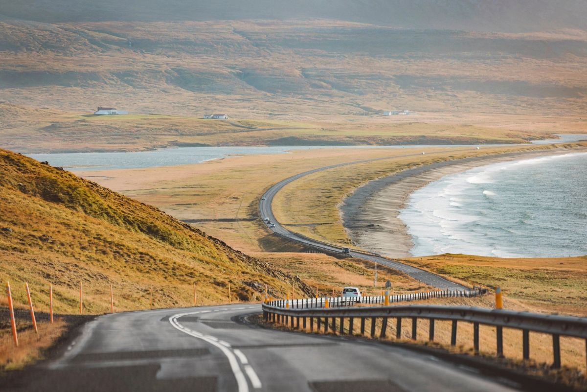 A paved road curving along a hillside above a bay with a pale beach and autumn-coloured hills