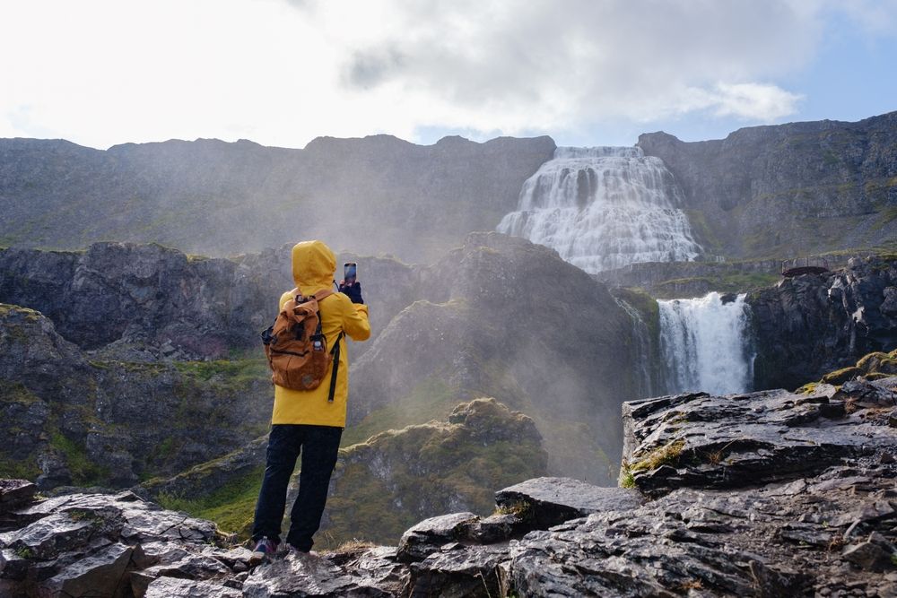 A traveler in a yellow rain jacket stands on rocky terrain, capturing the beauty of Dynjandi waterfall