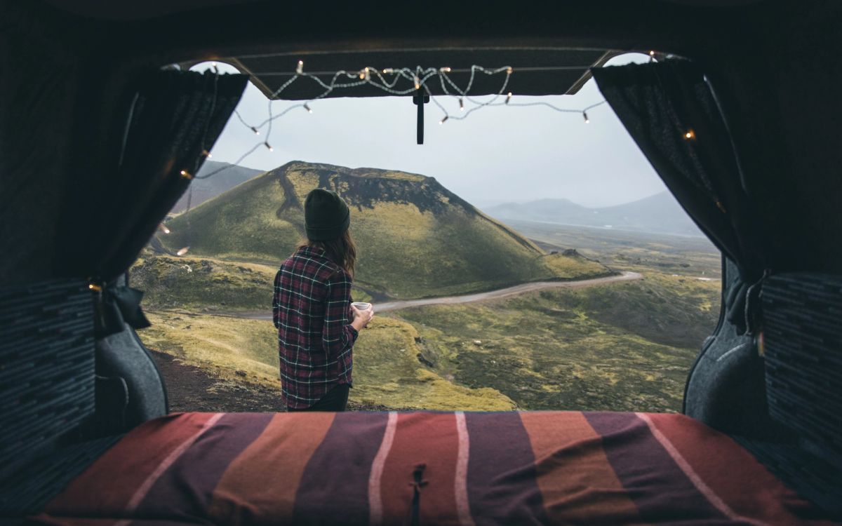 A campervan with open back doors parked on a hillside, a woman holding a mug and looking at a green volcanic crater
