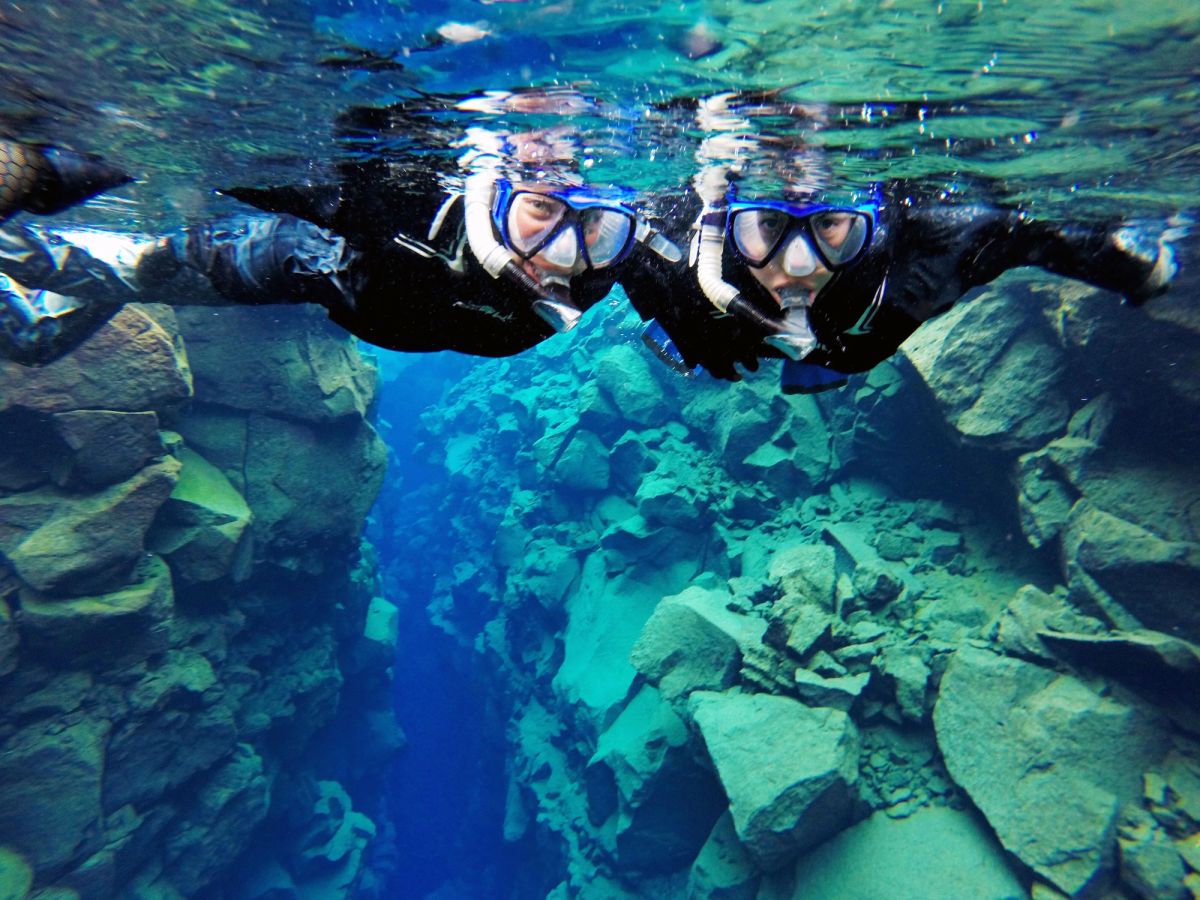 Two snorkellers in drysuits floating side by side above blue rocks in the clear water of Silfra