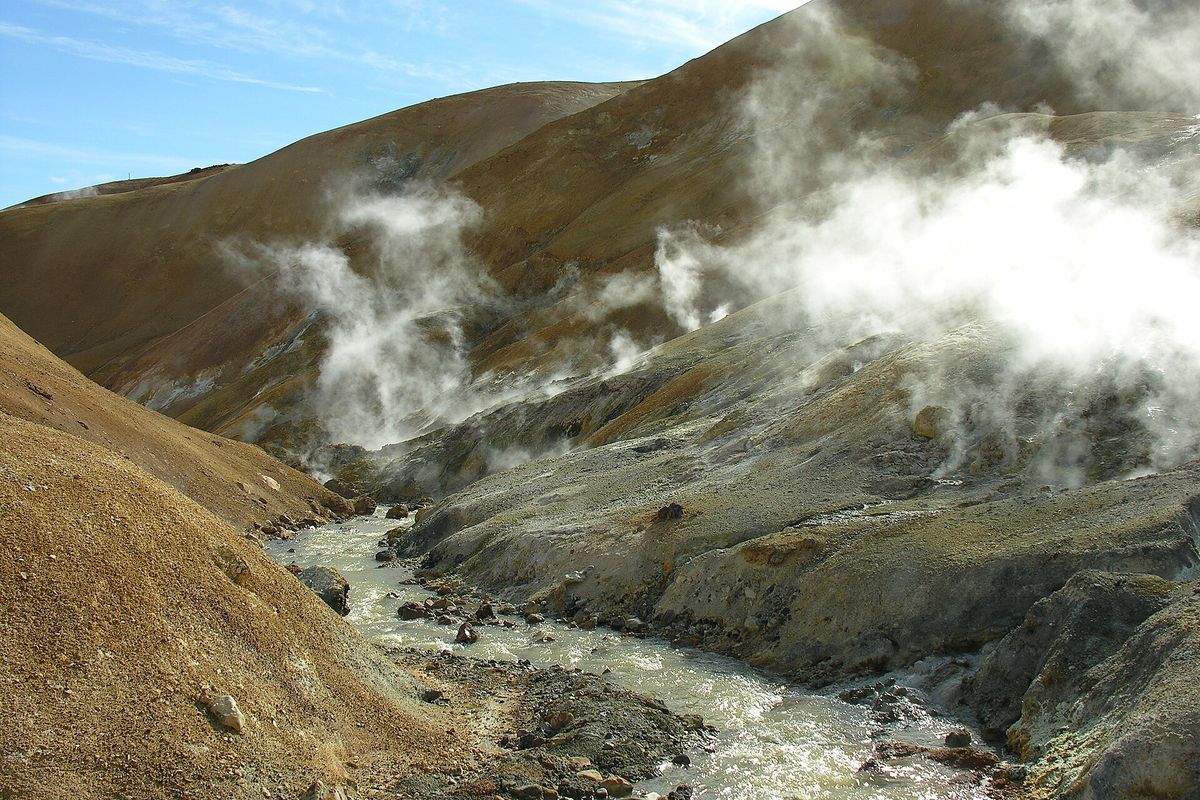 Steaming vents in a brown valley beside a fast stream, with green moss on the slopes