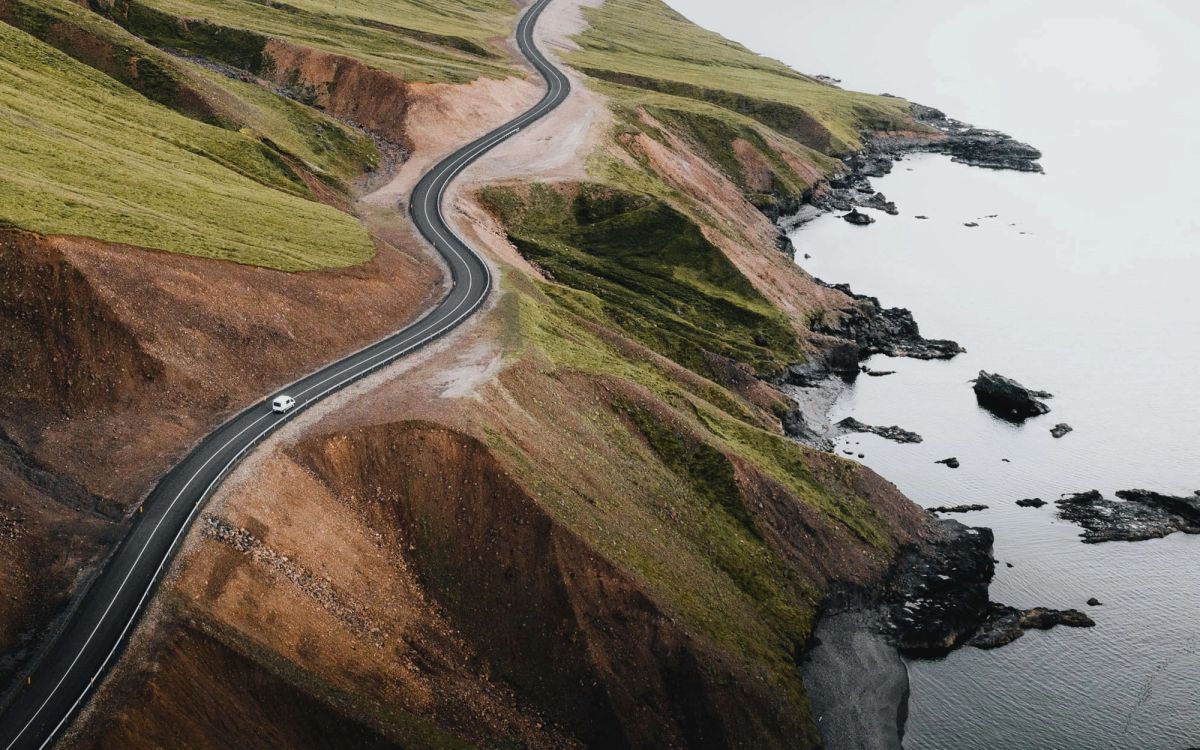 A road winding along a green coastal hillside above a rocky shore, seen from the air