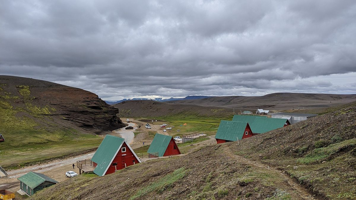 Red A-frame huts with green roofs on a slope above a valley, under a cloudy sky