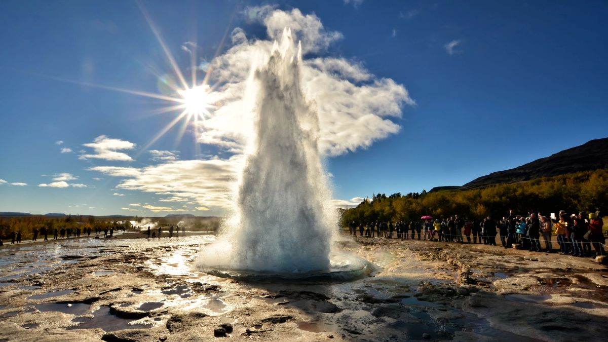 Strokkur geyser erupting in a tall white column under a bright sun, watched by a crowd