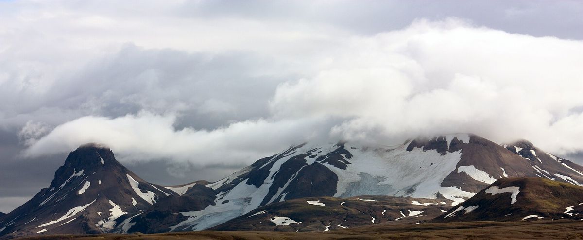 Dark, snow-capped mountains under a heavy band of cloud, seen across a flat plain