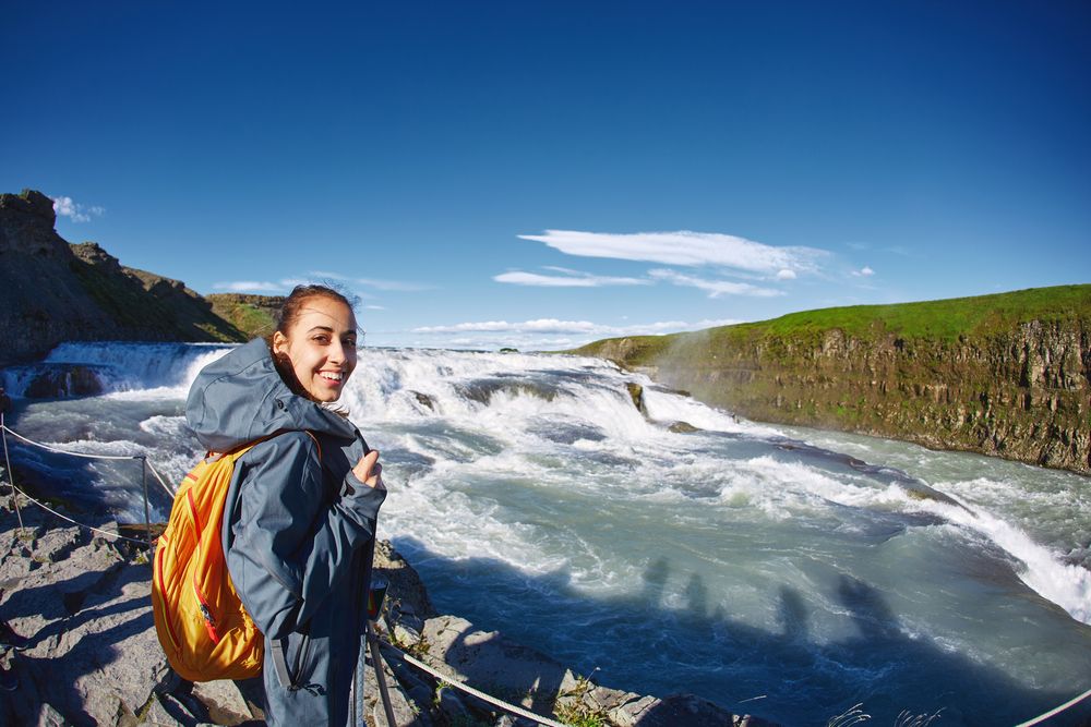 A girl in a waterproof jacket with a small orange backpack standing on a cliff overlooking Gullfoss Waterfall in Iceland