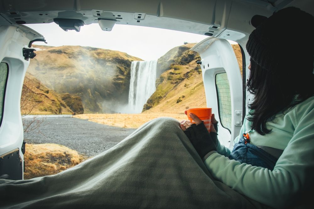 A tourist woman enjoying views of Skógafoss from the open door of a rented campervan