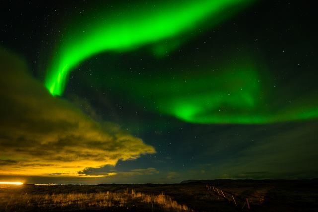 A green aurora ribbon over a bay with city lights on the horizon