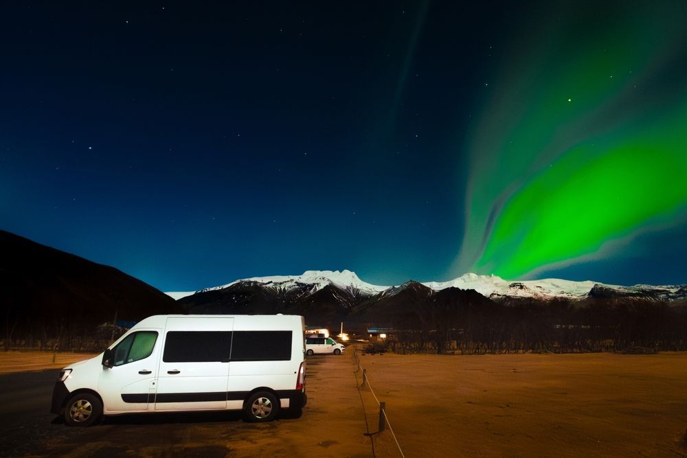 A white campervan parked under a band of green northern lights, with snowy mountains on the horizon