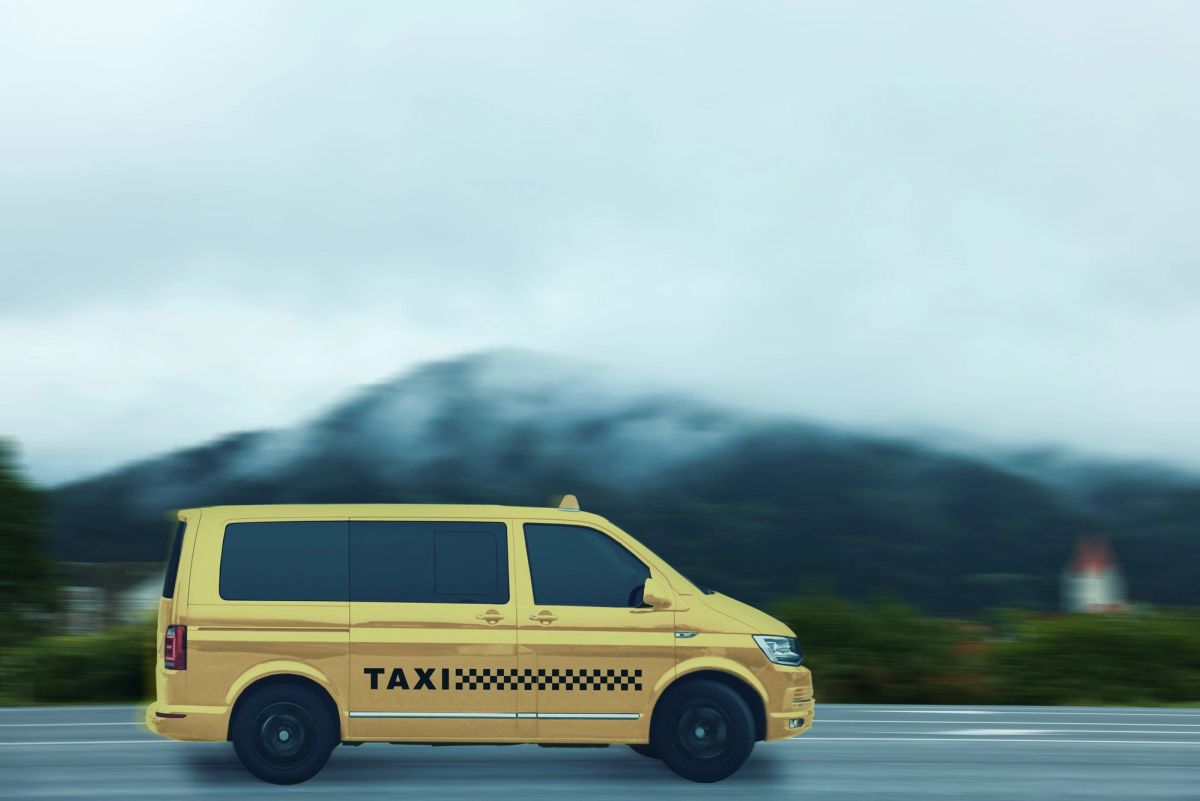 A yellow taxi van driving along a road with a cloud-covered mountain behind