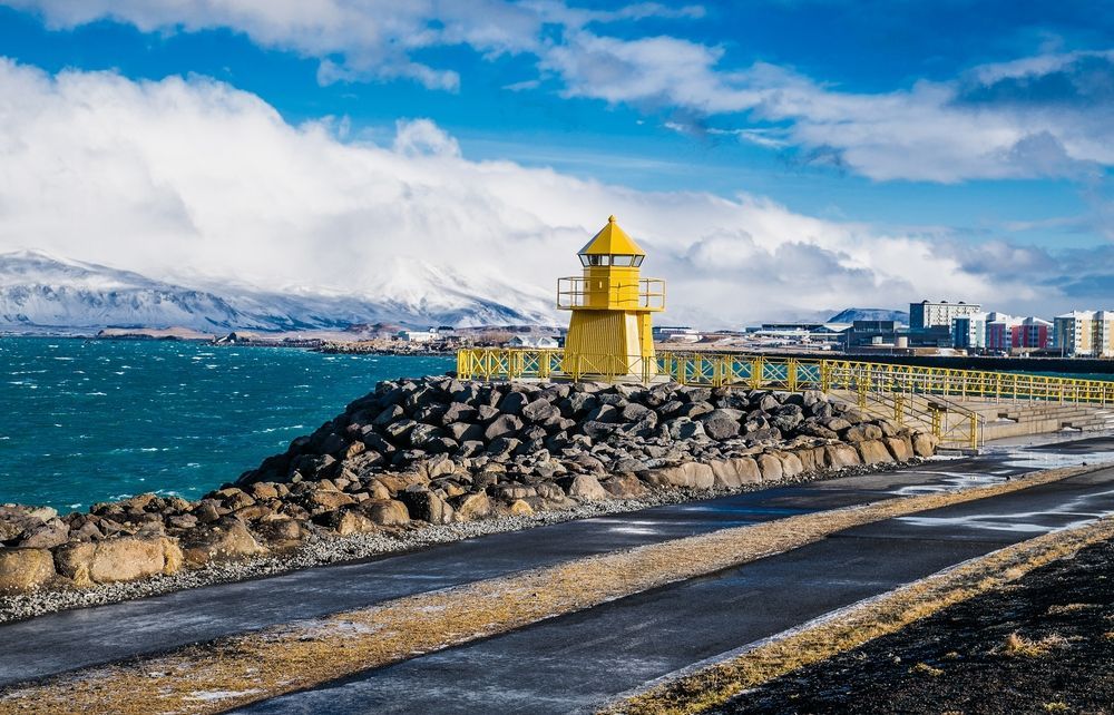 A small yellow lighthouse on a stone breakwater in Reykjavík, with snowy mountains across the water