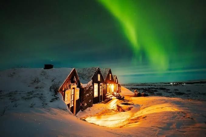 Turf-roofed cottages glowing with warm light under green northern lights on a snowy night