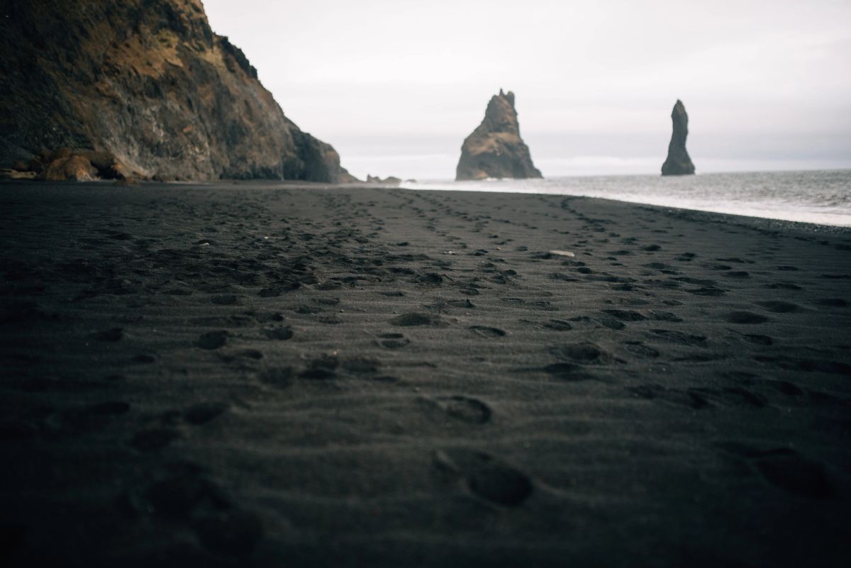 A black sand beach with footprints in the foreground and two dark sea stacks rising offshore