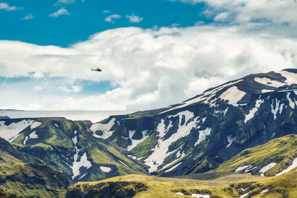 A small helicopter flying above green mountains with snow patches under a bright sky