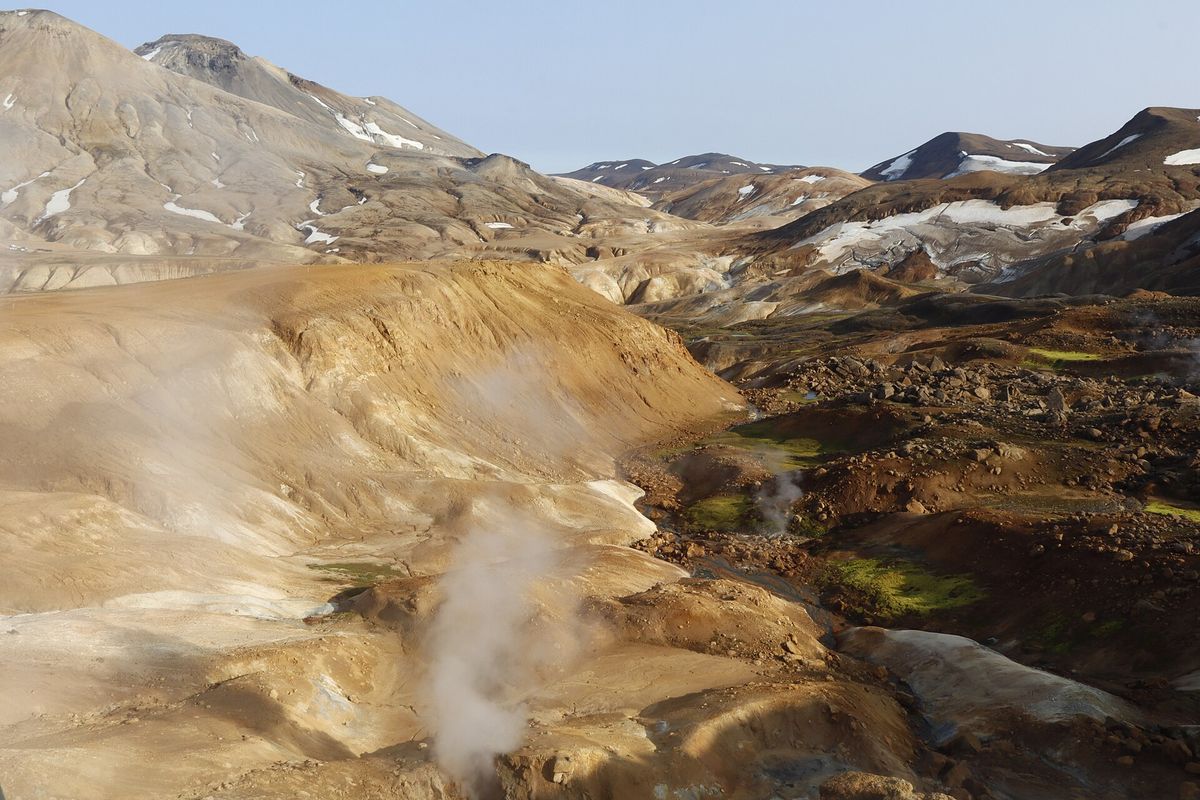 Steam rising from several vents across a valley of pale clay, with snow-patched mountains behind