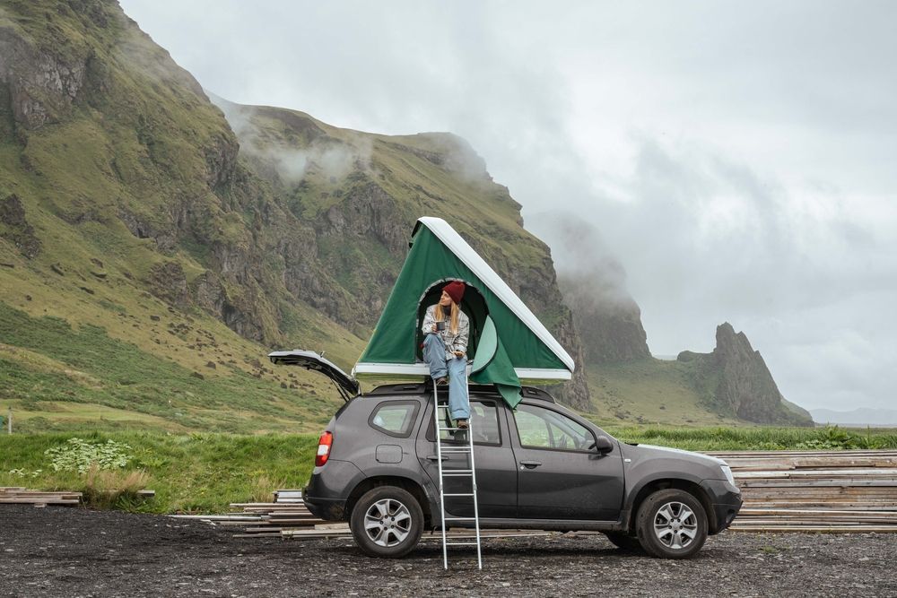 A person sitting in a rooftop tent on top of a 4x4 parked below steep green mountains