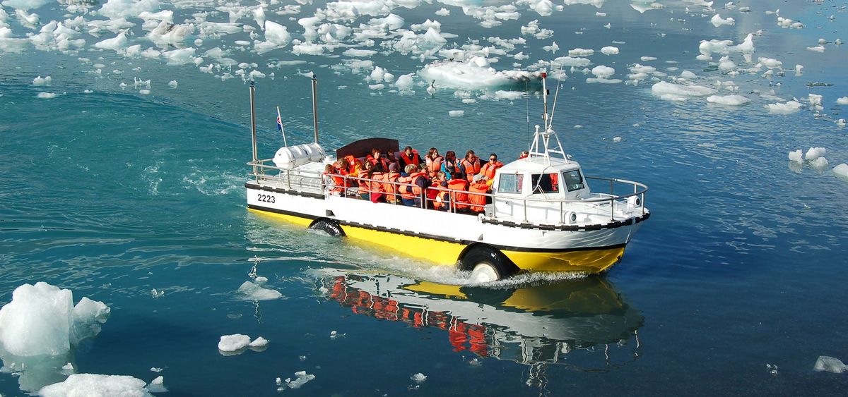 An amphibious boat full of passengers in orange life jackets sailing between icebergs at Jökulsárlón