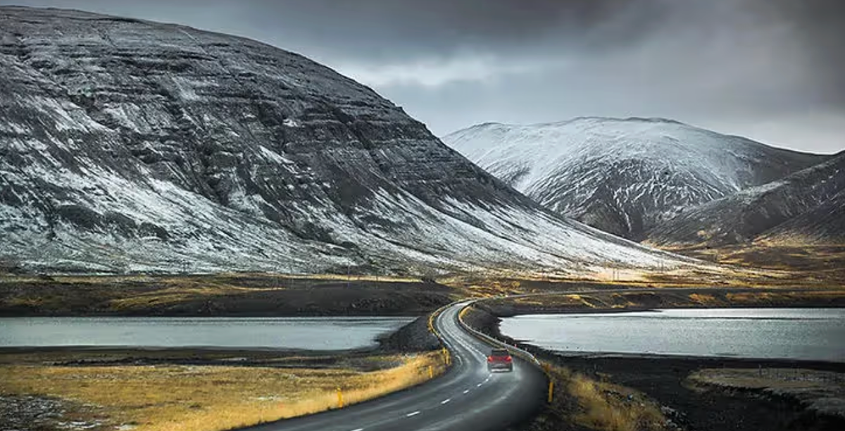 A car on a winding road between two lakes, with snow-dusted mountains rising behind