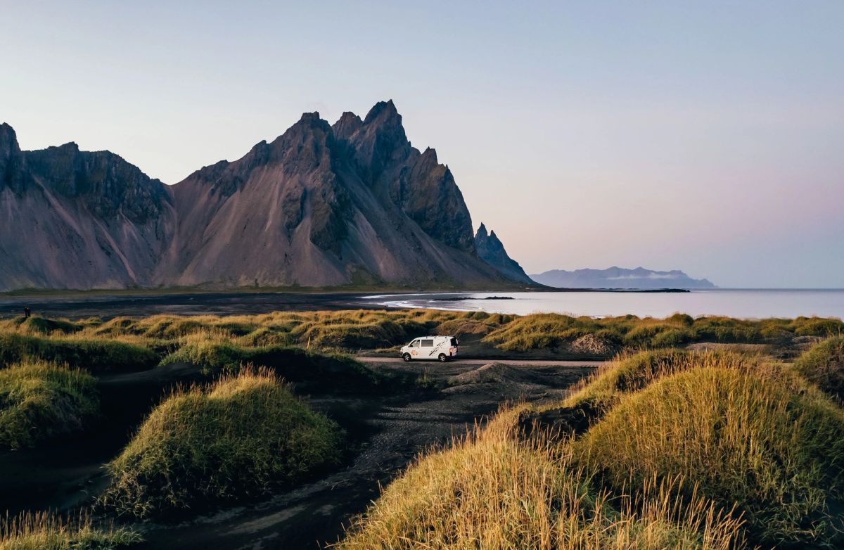 A white campervan parked on grassy dunes below jagged mountain peaks on a calm evening