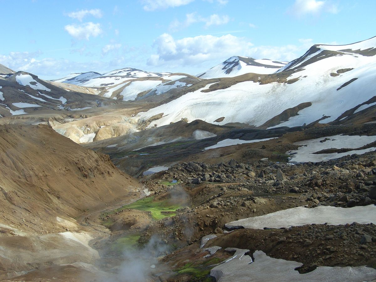 Snow-streaked mountains above a stony valley where steam rises from hot springs beside a stream