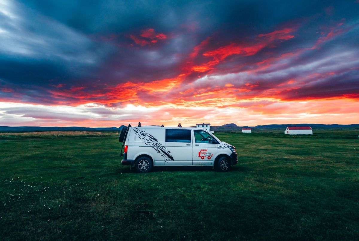 A white campervan parked in a green field under a fiery red and orange sunset sky