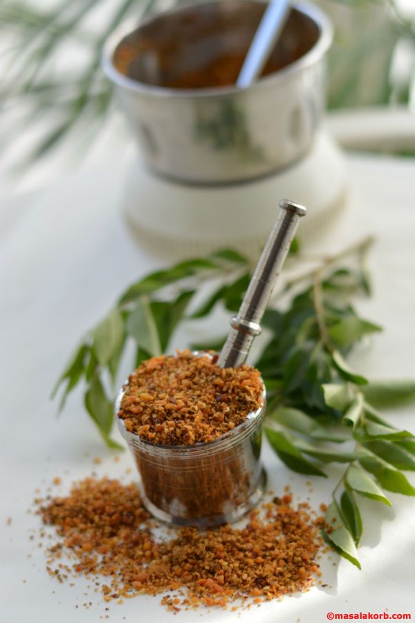 Coarse spice powder in a small glass with a steel spoon, curry leaves behind