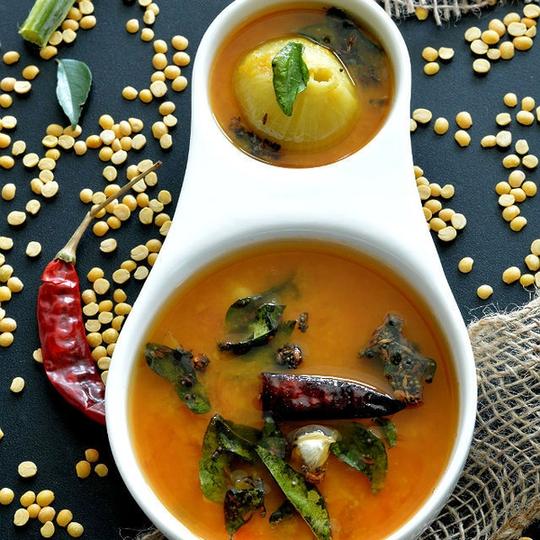 Pappu charu with curry leaves and a dry red chilli in a white bowl, toor dal scattered around
