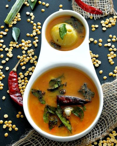 Pappu charu with curry leaves and a dry red chilli in a white bowl, toor dal scattered around