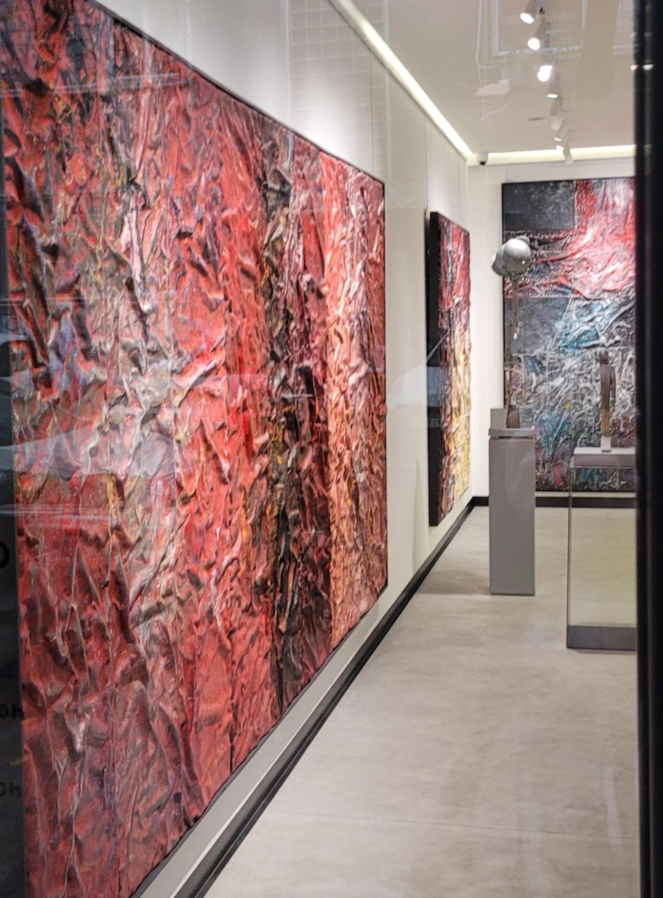 Gallery interior with large textured abstract paintings in shades of red, pink, and black displayed on white walls. The artworks have a wrinkled, three-dimensional surface. Sculptures on pedestals are visible further inside, under ceiling spotlights
