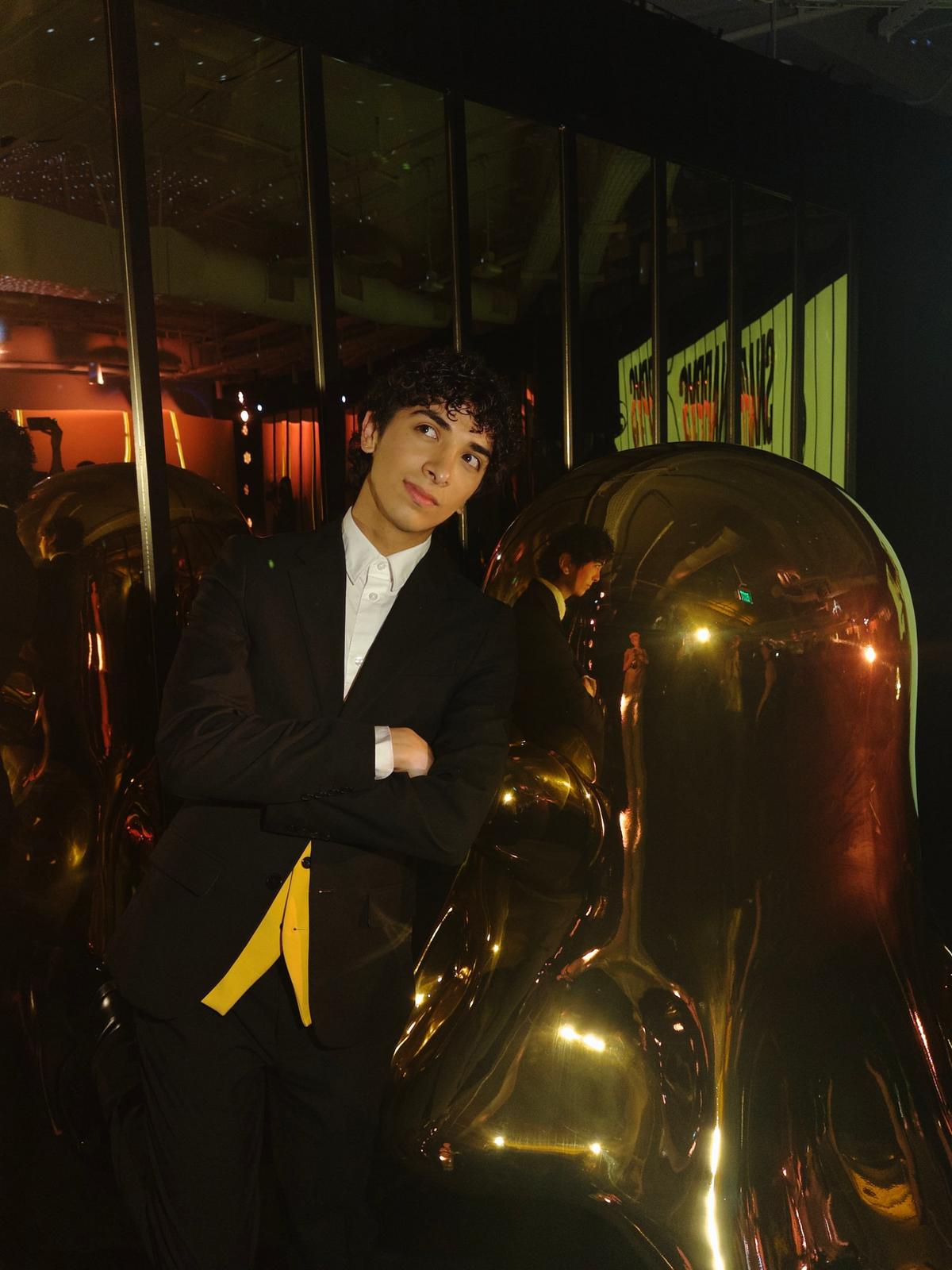 Joseph in a black suit leaning against a mirrored gold sculpture