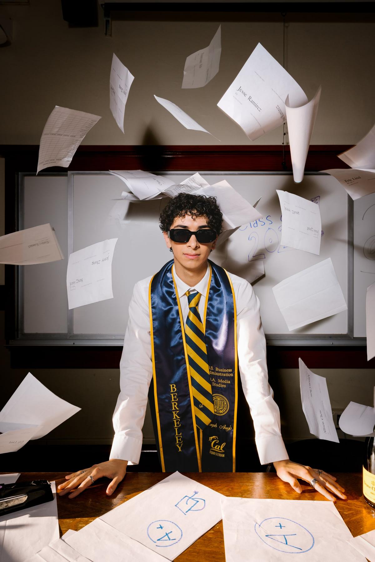 Joseph in sunglasses and a Berkeley graduation stole as papers fly around him
