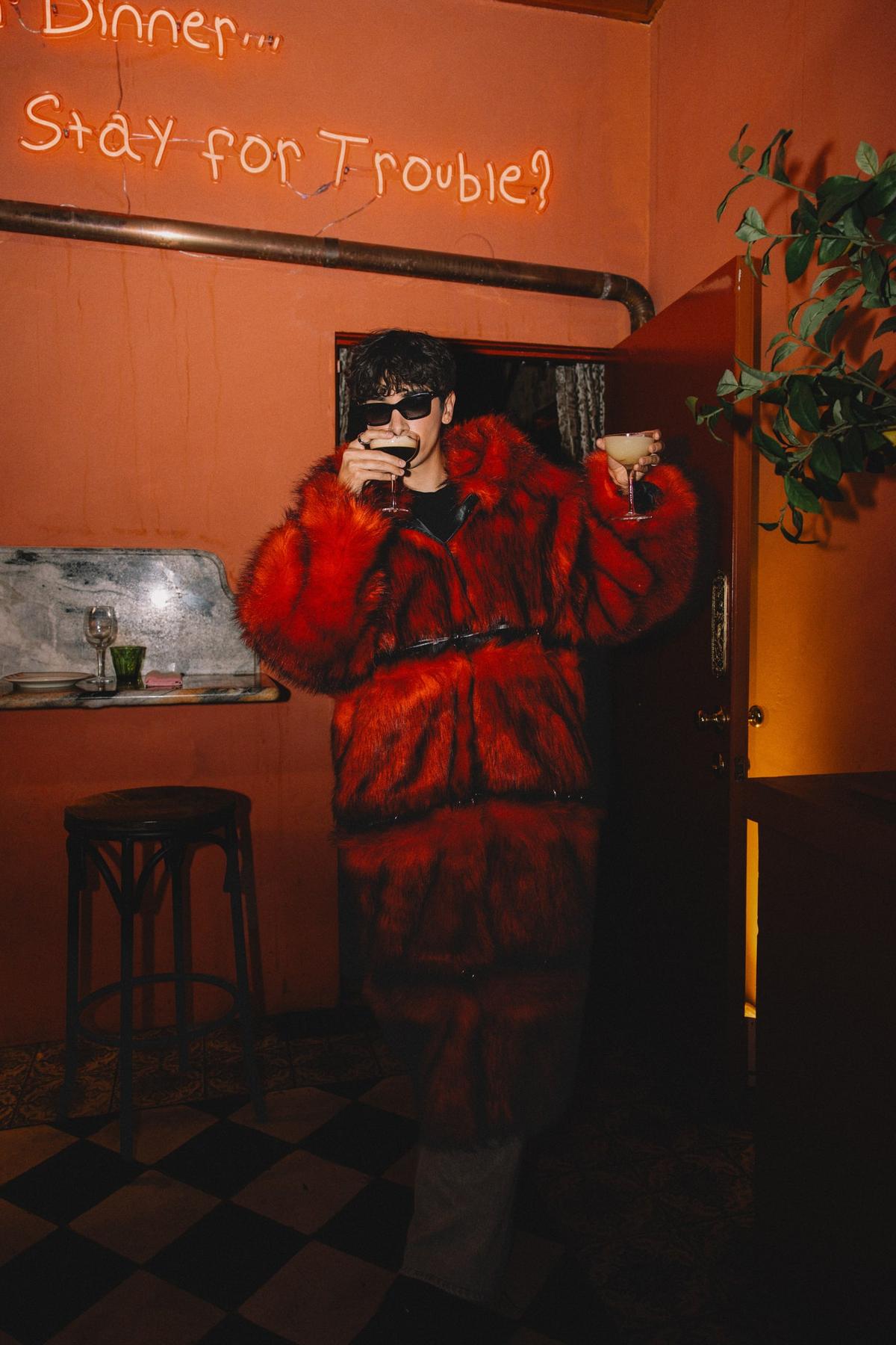 Joseph in a red faux-fur coat and sunglasses, holding two cocktails under a neon sign