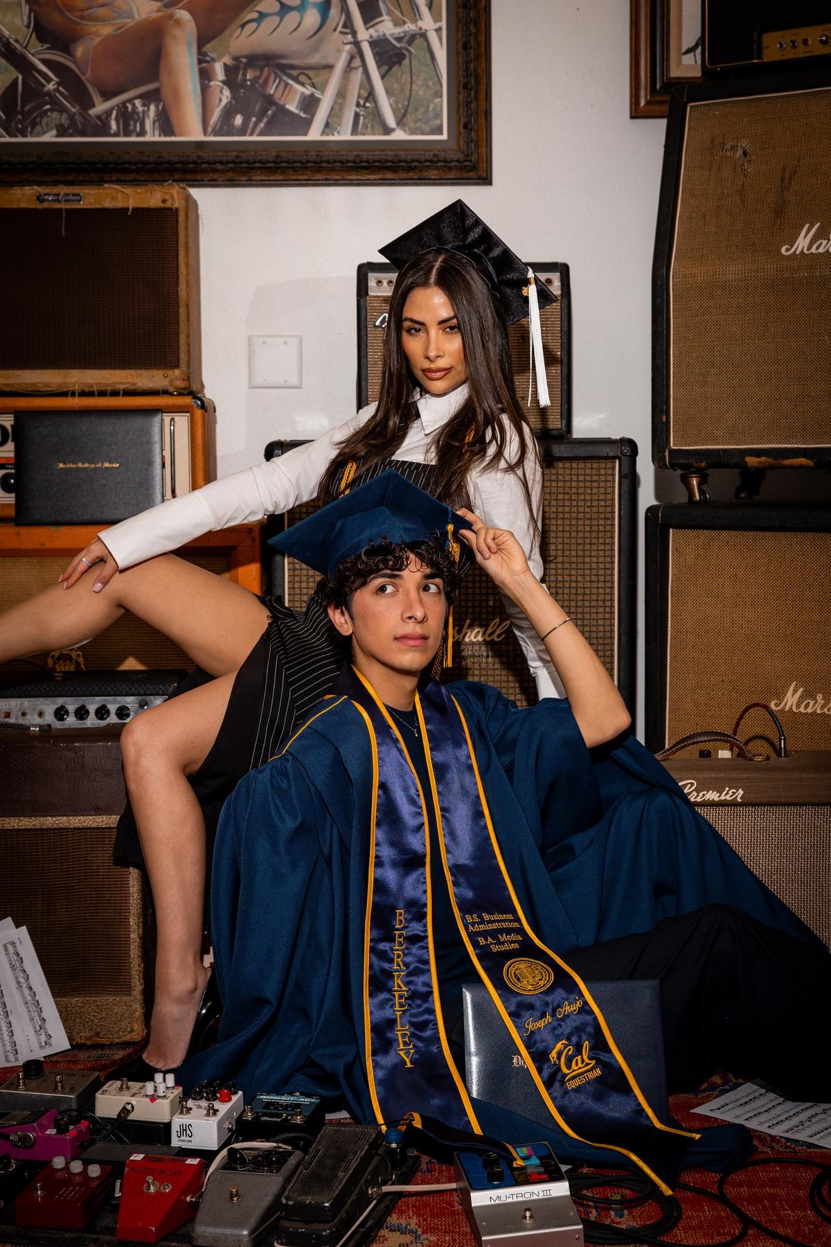 Joseph in a Berkeley graduation gown and cap, seated in front of guitar amplifiers with another graduate