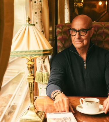 Stanley Tucci, wearing N.Peal knitwear, sits in a train carriage