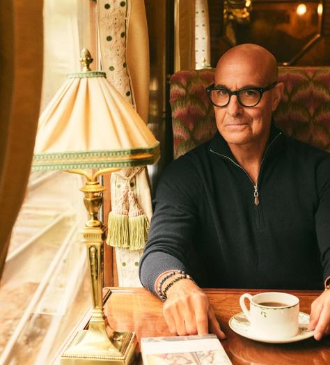 Stanley Tucci, wearing N.Peal knitwear, sits in a train carriage
