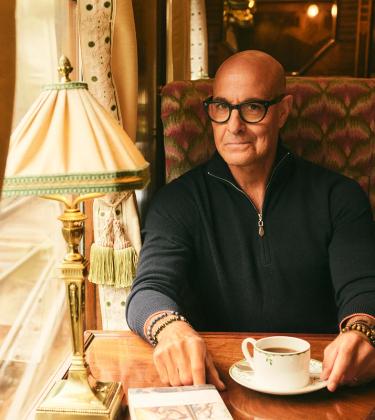 Stanley Tucci, wearing N.Peal knitwear, sits in a train carriage
