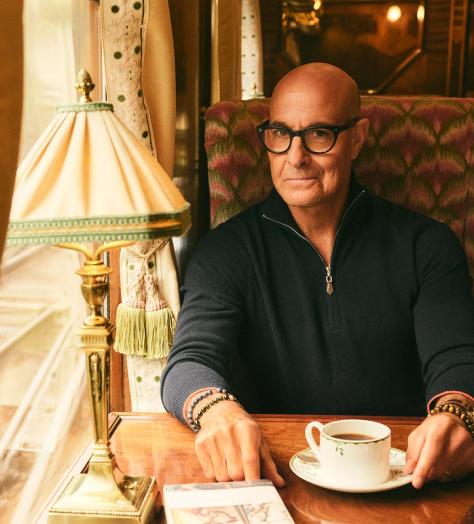 Stanley Tucci, wearing N.Peal knitwear, sits in a train carriage