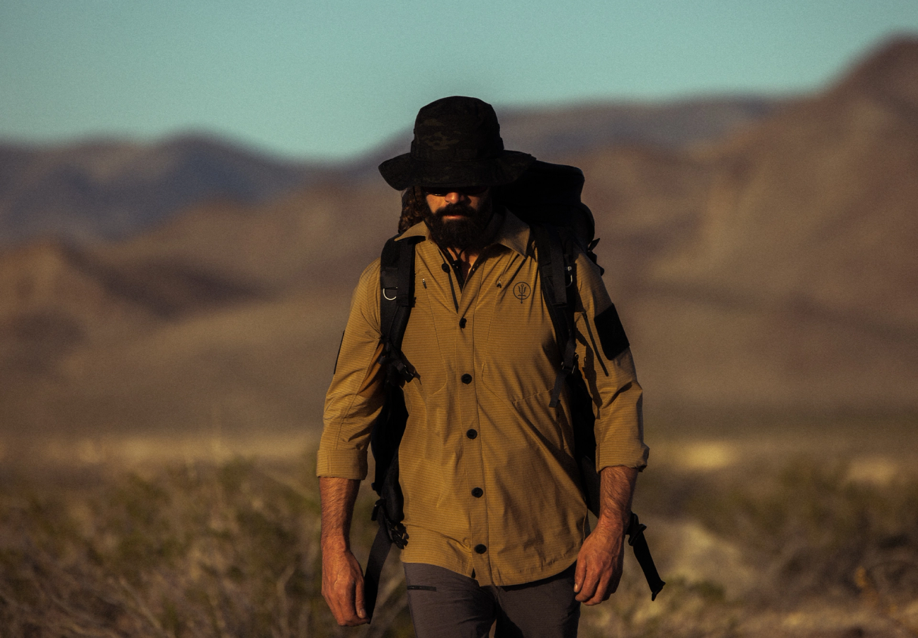 Man walking through dusty terrain wearing carbon shirt, watchman boonie hat and rucksack