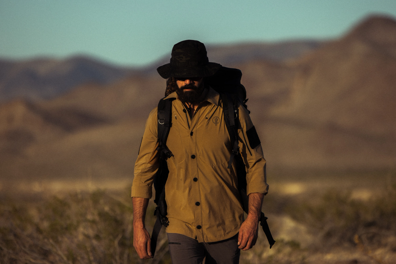 Man walking through dusty terrain wearing carbon shirt, watchman boonie hat and rucksack