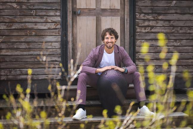 Nachson Mimran in a purple suit, white t-shirt and white trainers, sat on the steps outside a wooden hut
