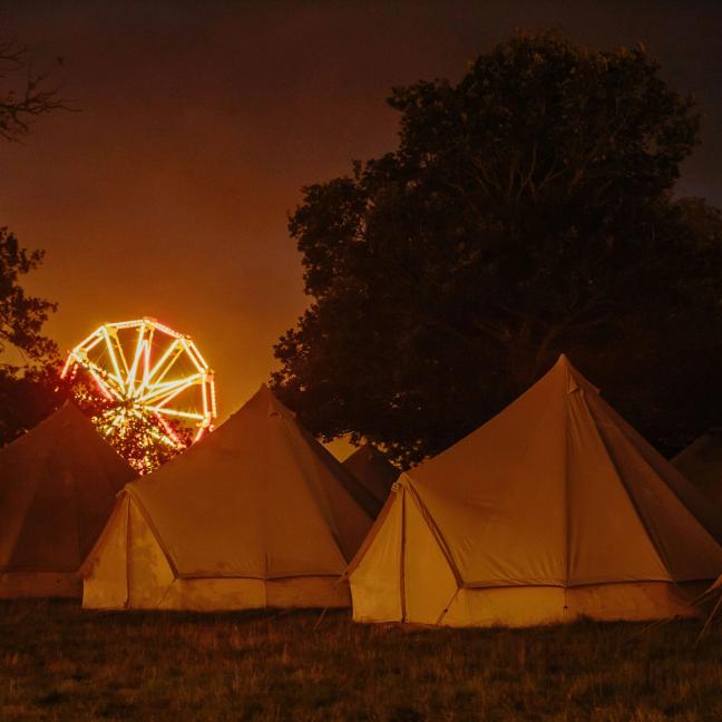 Pre-erected camping Tents in a field at night time with ferris wheel lit up in the background